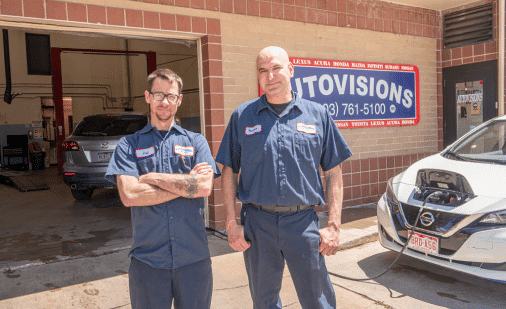 Two auto mechanics in blue uniforms stand outside the auto repair shop. One is crossing his arms while the other has his hands by his sides. They are in front of a brick building with a sign that reads "AUTOVISIONS" and lists various car brands serviced. A car is parked nearby.