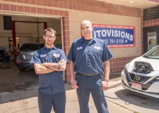 Two auto mechanics in blue uniforms stand outside the auto repair shop. One is crossing his arms while the other has his hands by his sides. They are in front of a brick building with a sign that reads "AUTOVISIONS" and lists various car brands serviced. A car is parked nearby.