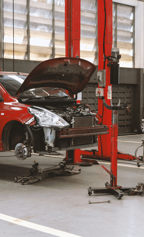 A red car with its hood open is lifted on a car lift in a garage, where the front bumper, wheels, and various parts are removed and placed around. Showcasing the meticulous work of skilled mechanics, this scene highlights a career dedicated to automotive excellence. The garage features red equipment and a concrete floor.