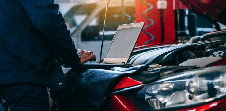 A mechanic stands next to a red car with its hood open, conducting a pre-purchase auto inspection while working on a laptop placed on the engine.