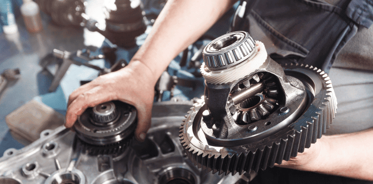 A person wearing a grey shirt is handling a gearbox component with intricate gears and bearings on a workbench. In the background, other mechanical parts and tools are visible, suggesting a workshop environment focused on machinery assembly or repair.