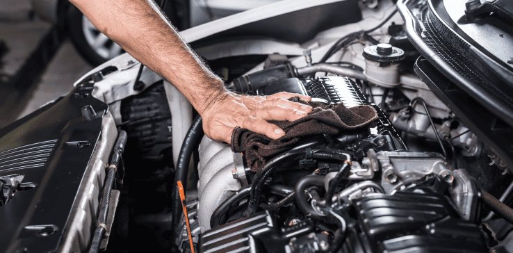 A person is engaged in vehicle maintenance, meticulously cleaning a car engine with a cloth. The open engine compartment reveals various components, hoses, and parts. The individual's arm is visible, showing delicate attention while wiping the surfaces to ensure the engine's cleanliness.