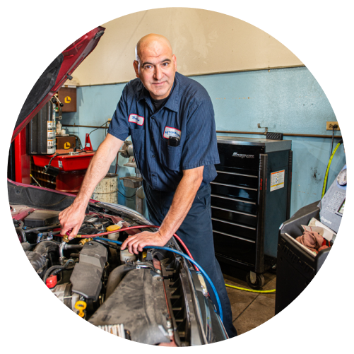 A man wearing a dark blue uniform works on the engine of a car in an auto repair shop. Tools and equipment are visible in the background.