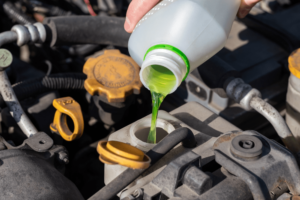 Essential car maintenance tips for winter prep in Englewood, CO. Image of mechanic hand pouring green colored antifreeze into the coolant reservoir on car in shop.