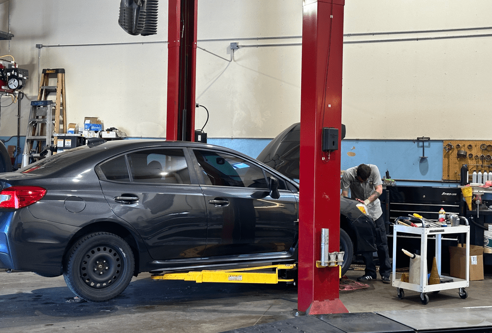 How much does a mechanic charge in Englewood, CO with Autovisions. Image of a car on a lift inside the shop for auto repairs and maintenance with mechanic working on engine under the hood.