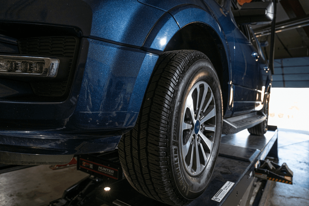 Colorado’s traction law, auto repair in Englewood, CO by Autovisions. A blue vehicle on a lift with a focus on its tires, ensuring accurate alignment for optimal driving performance and safety.