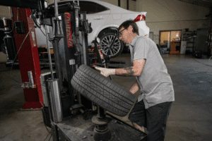Car maintenance, auto repair in Englewood, CO by Autovisions. Image of a technician mounting a tire using a tire machine with another vehicle lifted in the background. Highlighting Autovisions’ commitment to safe handling, proper installation, and reliable tire performance.