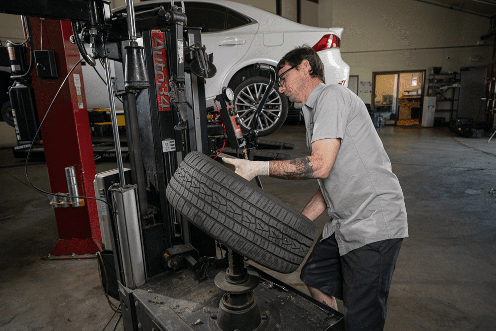 Car maintenance, auto repair in Englewood, CO by Autovisions. Image of a technician mounting a tire using a tire machine with another vehicle lifted in the background. Highlighting Autovisions’ commitment to safe handling, proper installation, and reliable tire performance.